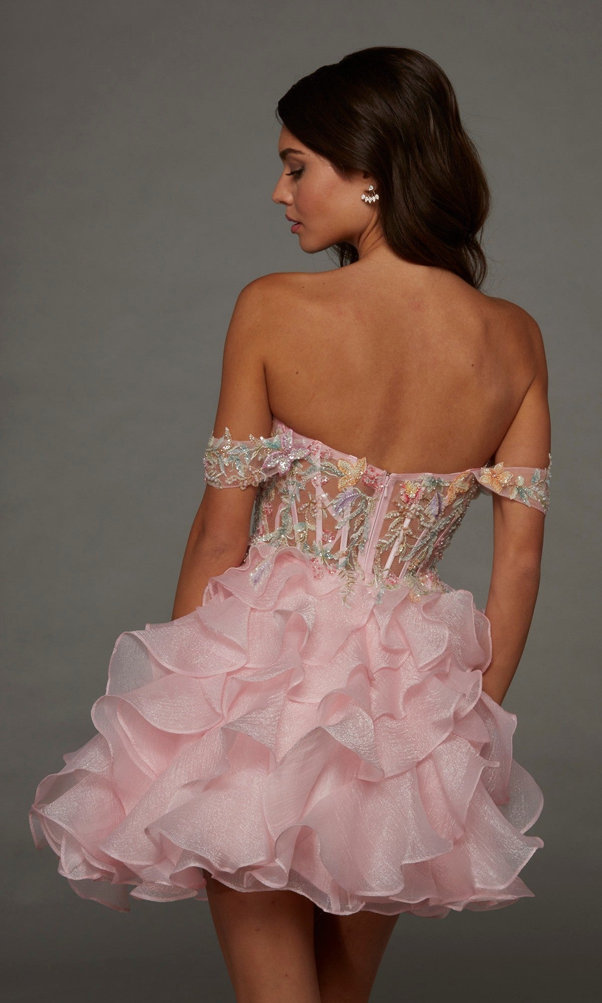 A woman wearing the Alyce 30087 Short Homecoming Dress—an off-shoulder, fit-and-flare pink mini dress featuring a ruffled skirt and beaded floral appliques on the bodice—stands facing away while glancing to the side against a plain background.